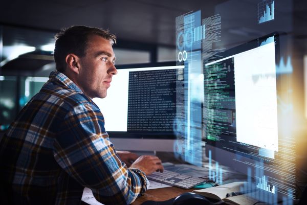 Man wearing a check shirt looking at a computer screen with dual screens
