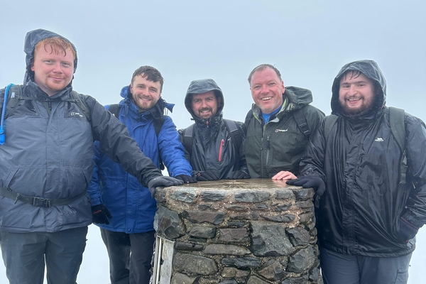 Five team members from Lucid Systems in waterproof jackets celebrate at Snowdon summit beside a stone cairn in misty, rainy weather.
